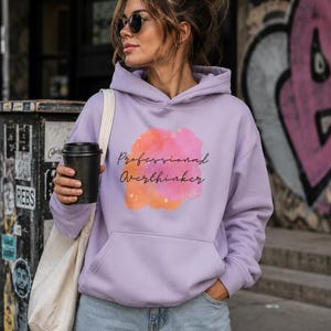 Könnte beinhalten: Ein helllila Kapuzenpullover mit einer Kängurutasche auf der Vorderseite und einem zentralen Grafikdesign. Die Grafik besteht aus einem rosa und orangefarbenen Aquarell-Fleck mit dem schwarzen kursiven Text Professional Overthinker.