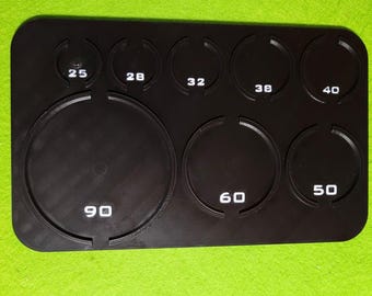 Può includere: Uno strumento di misurazione rettangolare in plastica nera con otto fori circolari di varie dimensioni. Ogni foro è contrassegnato da numeri bianchi stampati: 25, 28, 32, 38, 40, 50, 60 e 90, che indicano specifiche misure di diametro.