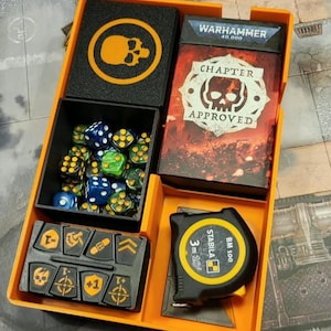 Könnte beinhalten: Ein orangefarbenes Kunststoff-Organizer-Tablett mit einer schwarzen Box mit Totenkopf-Symbol, einem Warhammer 40,000 Chapter Approved Kartendeck, einem schwarzen Behälter mit blauen und grünen Würfeln, einem schwarzen Token-Tablett und einem Stabila BM 100 3m Maßband.