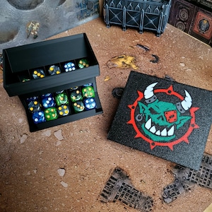 May include: A black tiered dice storage tray filled with multi-colored dice and a matching black lid featuring a graphic of a green orc face with white horns and a red eye patch surrounded by a red spiked circle design on the textured surface.