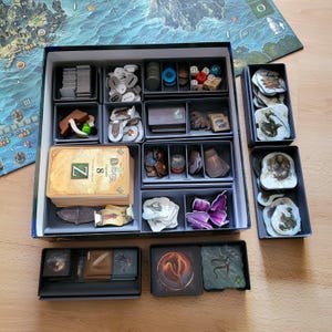 Könnte beinhalten: Eine Brettspielschachtel mit grauen Kunststoff-Organizern, gefüllt mit verschiedenen Spielkomponenten wie Kartenstapeln, kleinen Spielsteinen, Würfeln und illustrierten Pappteilen. Der Kartenstapel ist mit dem Buchstaben Z und der Zahl 8 beschriftet.