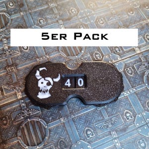 Könnte beinhalten: Ein schwarzer, texturierter Spielzähler mit einem weißen Totenkopf-Symbol mit Hörnern und einem Auge, der die Zahl 40 auf einem mechanischen Zifferblatt anzeigt. Das Gerät ist mit dem Text 5ER PACK beschriftet, was ein Set von fünf Einheiten anzeigt.