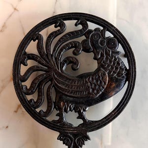 Könnte beinhalten: Schwarzer gusseiserner Topfuntersetzer mit detailliertem Hahn-Design in einem kreisförmigen Rahmen. Der Topfuntersetzer hat einen verzierten Griff und schützt Oberflächen vor heißem Kochgeschirr. Die Schwanzfedern des Hahns sind deutlich zu sehen.