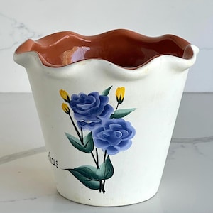 Peut inclure: Un pot de fleurs en céramique blanche avec un bord ondulé et festonné et un intérieur couleur terre cuite. L'extérieur présente un motif peint à la main de deux roses bleues avec des feuilles vertes et de petits boutons jaunes.