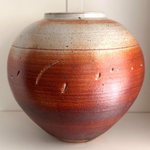 Könnte beinhalten: Eine handgefertigte Keramikvase mit rundem Körper und breiter Öffnung. Der obere Teil ist gesprenkeltes Off-White, das in einen satten, rotbraunen Farbton übergeht. Die Vase weist geschnitzte Details und eine strukturierte Oberfläche auf, die ihre handwerkliche Qualität hervorhebt.