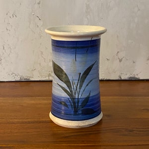 Puede incluir: Un jarrón de cerámica blanca con un diseño floral azul y blanco. El jarrón tiene una forma cilíndrica y un borde ligeramente acampanado.