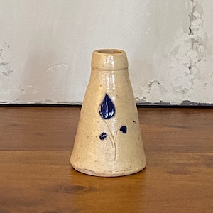 Peut inclure: Un petit vase en céramique de couleur crème avec un motif floral bleu. Le vase est en forme de cône et a une ouverture étroite.