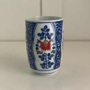 May include: A white ceramic teacup with a blue and white floral design. The cup features a red flower with blue and green leaves.