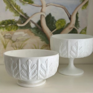 Puede incluir: Dos cuencos de cerámica blanca con un diseño de hojas. Los cuencos están sobre una superficie blanca delante de una pintura de un árbol.