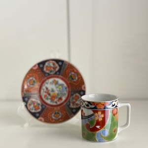 Puede incluir: Un juego de tazas y platillos de té de estilo japonés con un diseño floral y geométrico colorido. La taza y el platillo son ambos blancos con acentos rojos, azules, verdes y dorados.