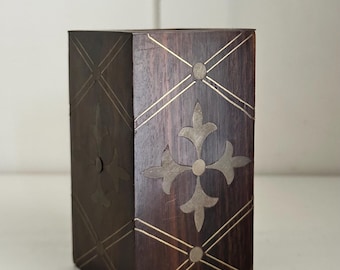 May include: A dark brown wooden box with a geometric design. The box features inlaid metal accents in a cross and diamond pattern. The box is rectangular and has a decorative, vintage aesthetic.