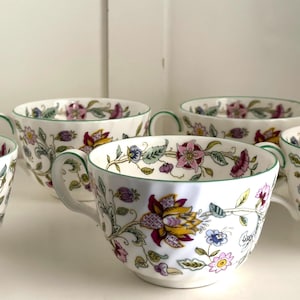 May include: A collection of white teacups with floral patterns in pink, yellow, and blue. Each cup has a green handle and a green trim along the rim. The cups are arranged on a white surface.