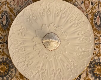 Könnte beinhalten: Weiße Keramik-Servierplatte mit goldfarbenem Rand und einem verzierten Design. Die Platte hat einen zentralen Griff mit einem roten Akzent.