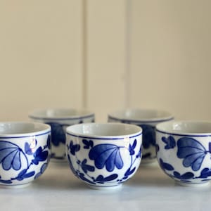 Pode incluir: Cinco pequenas chávenas de cerâmica branca com um desenho floral azul. As chávenas estão dispostas em fila, mostrando os intrincados padrões azuis sobre o fundo branco. As chávenas são provavelmente para chá ou saquê.