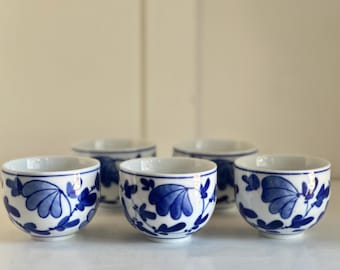 Könnte beinhalten: Fünf kleine, weiße Keramikbecher mit blauem Blumenmuster. Die Becher sind in einer Reihe angeordnet und zeigen die komplizierten blauen Muster vor dem weißen Hintergrund. Die Becher sind wahrscheinlich für Tee oder Sake.
