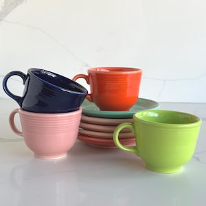 May include: A collection of ceramic cups and saucers in various colors including navy blue, light pink, bright orange, lime green, and soft teal. The cups feature a rounded shape with a side handle and horizontal ribbed detailing near the rim.