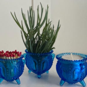 May include: Three blue glass candle holders with a decorative design. The center holder has a small white candle inside. The holder on the left has a pile of red matchsticks inside. The holder on the right is empty.
