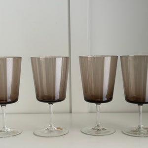 May include: Four clear glass wine glasses with a dark brown tint. The glasses are arranged in a row on a white surface.