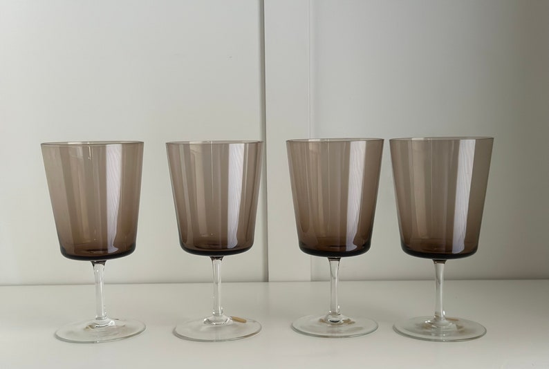 May include: Four clear glass wine glasses with a dark brown tint. The glasses are arranged in a row on a white surface.