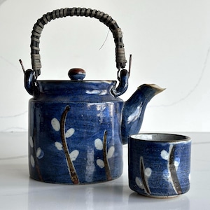 May include: A blue ceramic teapot and matching cup set. The teapot has a woven handle and a small lid. Both pieces are decorated with white floral designs on a dark blue background. The teapot has a spout for pouring.