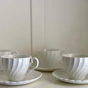 Puede incluir: Cuatro tazas de té blancas y platillos con un diseño de espiral. Las tazas y los platillos están dispuestos en una fila sobre una superficie blanca.