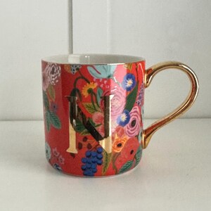 Può includere: Una tazza in ceramica rossa con manico dorato e la lettera "N" in oro. La tazza è decorata con un motivo floreale in vari colori, tra cui rosa, blu e giallo. La tazza è alta circa 10 cm.