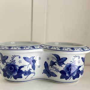 May include: A white ceramic planter with two connected compartments. The planter is decorated with blue floral and butterfly designs. The rim has a blue floral pattern. The planter is likely used for flowers or small plants.