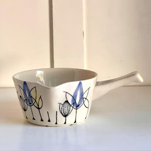 Puede incluir: Un pequeño cuenco de cerámica blanquecina con asa, decorado con diseños florales azules, amarillos y negros. El cuenco tiene un borde curvo y un pico vertedor. Una adición encantadora a cualquier cocina.