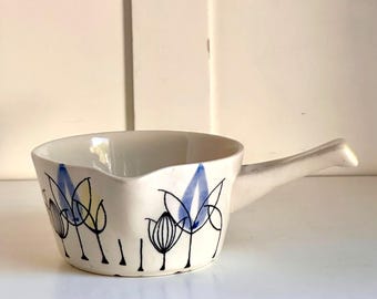 May include: A small, off-white ceramic bowl with a handle, decorated with blue, yellow, and black floral designs. The bowl has a curved rim and a spout for easy pouring. A charming addition to any kitchen.