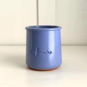 Op de afbeelding: Een kleine, licht periwinkle-kleurige keramische pot met een licht uitlopende rand. De woorden "la fermière" zijn in reliëf op de voorkant. De basis van de pot is terracotta van kleur.