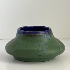 May include: A small, handcrafted ceramic vase with a unique shape. The vase has a green exterior with a textured surface and a dark blue interior. The rim is accented with a blue glaze. The vase has a wide, rounded base and a flared top.