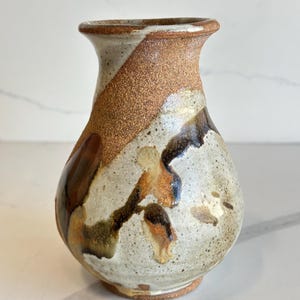 May include: A handmade ceramic vase with a speckled, off-white base. The vase features a textured, brown diagonal stripe and abstract brown and orange accents. The vase has a rounded body and a slightly flared rim, ideal for displaying flowers.