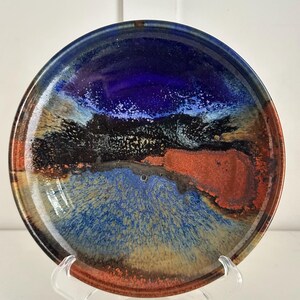 May include: A round ceramic plate with a glossy finish. The plate features a vibrant abstract design with shades of blue, black, orange, and brown. The plate is displayed on a clear plastic stand, showcasing its artistic and decorative nature.