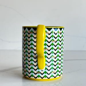Op de afbeelding: Een witte keramische mok met een zwarte handgreep en de tekst "But First, Coffee" in een vetgedrukte, zwarte letter. De mok staat tegen een vage achtergrond, wat een gezellige setting suggereert.