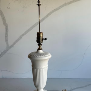 Peut inclure: Une lampe de table en céramique blanche avec une base en forme de vase cannelé et une base carrée de couleur laiton. La lampe a une longue tige en laiton et une ampoule ovale vert clair. Le cordon de la lampe est visible.