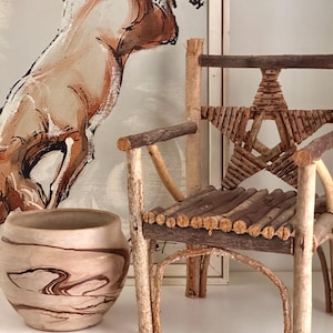 Puede incluir: Una silla rústica hecha a mano con ramas naturales, con un respaldo en forma de estrella. El asiento y el respaldo están construidos con pequeñas ramas horizontales. Un jarrón decorativo de cerámica veteada está en primer plano, con una pintura de un caballo en el fondo.