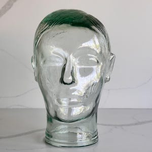Könnte beinhalten: Eine klare Glaskopfskulptur mit grün getöntem Haar. Die Skulptur hat ein detailliertes Gesicht mit Nase, Augen und Ohren. Der Kopf wird auf einer weißen Oberfläche ausgestellt.