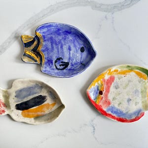 Puede incluir: Tres platos de cerámica pintados a mano con forma de pez. Uno es azul con una aleta amarilla y negra, otro es blanco con pinceladas azules, negras y naranjas, y el tercero es blanco con rayas rojas, amarillas, verdes y azules.