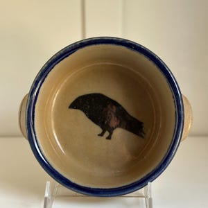 Puede incluir: Un cuenco pequeño y redondo de cerámica con interior beige y borde azul oscuro. El cuenco presenta una ilustración de un cuervo negro en el centro. Dos asas pequeñas a cada lado. El cuenco se exhibe sobre un soporte transparente.