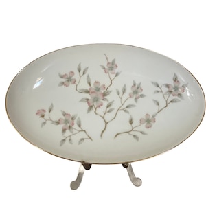 May include: A white oval serving dish with a gold rim and a floral design featuring pink and green flowers. The dish is on a metal stand with three legs.