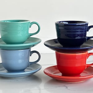 May include: Four ceramic tea cups and matching saucers arranged in two stacks. The left stack features a teal cup over a light blue cup. The right stack features a dark navy blue cup over a bright red cup. Each piece has concentric ring details.