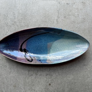 Puede incluir: Un plato de servir ovalado de cerámica largo con un esmalte azul, morado y blanco. El plato tiene un diseño abstracto negro pintado en la superficie.