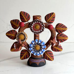 Könnte beinhalten: Eine Terrakotta-Skulptur in Form eines Lebensbaums mit herzförmigem Rahmen, handbemalten Blumen in Blau, Weiß und Braun, zwei kleinen Vögeln und mehreren gestreiften Blättern. Der Sockel ist schwarz mit einer violetten Wellenlinie.