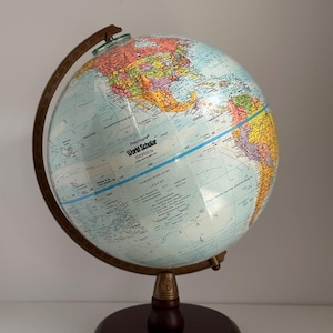 Może przedstawiać: Globus Replogle World Scholar Series z kolorową mapą świata. Globus jest zamontowany na ciemnobrązowej drewnianej podstawie z mosiężnym południkiem i wspornikiem. Globus przedstawia niebieski ocean i legendę.
