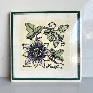 May include: A square ceramic tile featuring a botanical illustration of a Passiflora flower with light purple petals, dark purple filaments, and green leaves. The design includes the text Passiflora and a small copyright symbol in the corner.