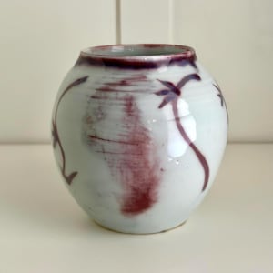 May include: A small, round ceramic vase with a white base and a dark burgundy rim. The vase features abstract burgundy floral designs. The vase has a smooth, glossy finish and is suitable for displaying small flowers or as a decorative accent.