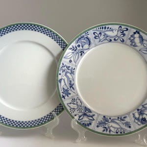 May include: Two white ceramic plates with blue and green decorative borders. One plate has a blue and white checkered border, while the other features a floral and bird design. Both plates are displayed on clear stands.