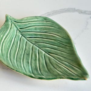 May include: A ceramic trinket dish shaped like a leaf with a glossy light green glaze. The surface features raised, textured veins that mimic the natural structure of a leaf, with darker green shading in the crevices and a pointed tip.