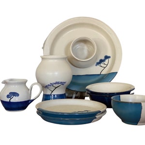 May include: A set of white ceramic dishes with blue accents. The set includes a large serving dish, two bowls, a jug, a vase, and two smaller plates. The dishes are decorated with a simple blue tree design.