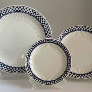 Puede incluir: Un juego de tres platos de cerámica blanca con un diseño de borde azul en forma de trébol. Los platos son de diferentes tamaños y se exhiben en soportes de plástico transparente. Los platos tienen una textura acanalada.